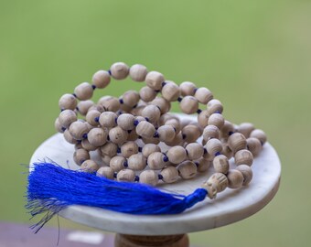 Könnte beinhalten: Eine Mala-Halskette aus Holzperlen mit einem leuchtend blauen Quasten, die auf einer weißen Marmoroberfläche liegt. Die Perlen sind hellbraun und mit dunkelblauem Faden aufgefädelt. Der Hintergrund ist ein sanftes Grün.