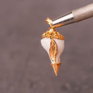 以下が含まれることがあります： 白い貝殻のデザインの金色のペンダント。ペンダントは、灰色の背景に対して銀色のツールで保持されています。貝殻には金のアクセントと装飾的な金のキャップが付いています。