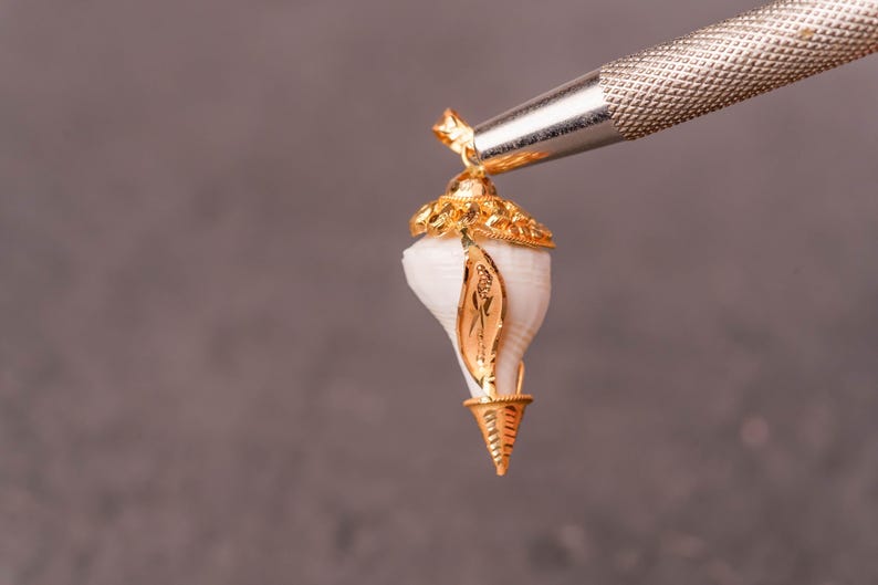以下が含まれることがあります： 白い貝殻のデザインの金色のペンダント。ペンダントは、灰色の背景に対して銀色のツールで保持されています。貝殻には金のアクセントと装飾的な金のキャップが付いています。