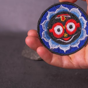 Op de afbeelding: Ronde geborduurde patch met een lachend gezicht. Het gezicht heeft grote rode ogen, een gele neus en een rode mond, tegen een zwarte achtergrond. Het is omgeven door een blauw en wit bloemenpatroon.