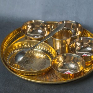 Puede incluir: Un juego de thali de latón dorado con múltiples cuencos pequeños, una taza central, un plato pequeño y una cuchara. El thali tiene un diseño grabado y ornamentado. El juego está diseñado para servir comida.