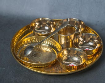 May include: A golden brass thali set with multiple small bowls, a central cup, a small plate, and a spoon. The thali has an ornate, engraved design. The set is designed for serving food.