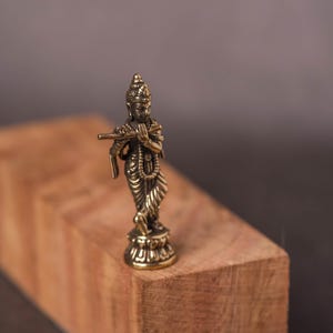 May include: A small, detailed brass statue of a deity playing a flute. The figure is standing on a decorative base and is set against a blurred wooden block and dark background. The statue is approximately 3 inches tall.