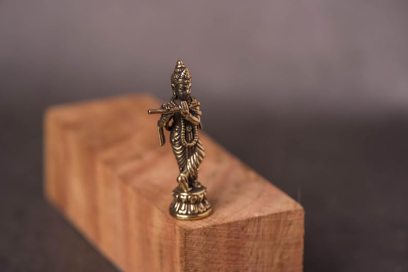 May include: A small, detailed brass statue of a deity playing a flute. The figure is standing on a decorative base and is set against a blurred wooden block and dark background. The statue is approximately 3 inches tall.