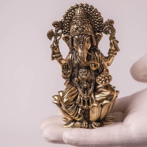 May include: A detailed brass statue of the Hindu deity Ganesha, seated on a lotus flower. The statue features intricate carvings, a large head, and multiple arms holding various objects. The figure is adorned with jewelry and a decorative headdress.