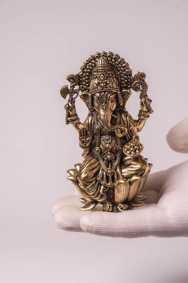 May include: A detailed brass statue of the Hindu deity Ganesha, seated on a lotus flower. The statue features intricate carvings, a large head, and multiple arms holding various objects. The figure is adorned with jewelry and a decorative headdress.