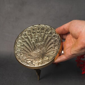 May include: A gold-colored, round decorative object with a peacock design. The peacock's feathers are fanned out, and the object is held by a hand. The object is on a dark surface.