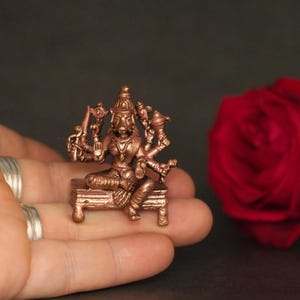 May include: A small, copper-colored statue of a multi-armed deity seated on a bench. The statue has intricate details and is held in a hand. A red rose is in the background. The statue is likely a religious or decorative item.