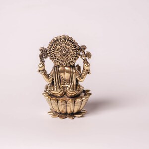 May include: A golden-colored statue of a deity, viewed from the back. The figure is seated on a lotus flower base. The statue has intricate details, including a halo and multiple arms holding objects. The overall design is ornate and detailed.