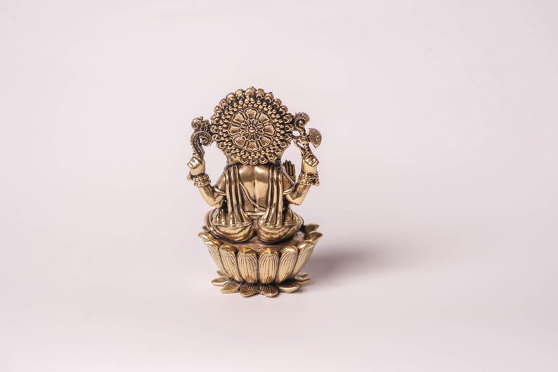 May include: A golden-colored statue of a deity, viewed from the back. The figure is seated on a lotus flower base. The statue has intricate details, including a halo and multiple arms holding objects. The overall design is ornate and detailed.