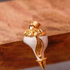 以下が含まれることがあります： 金色の巻貝の形をしたペンダント。ペンダントは、上部と貝殻の表面に沿って複雑な金のディテールが施されています。貝殻は白で、金のアクセントとチェーン用の小さなループが付いています。