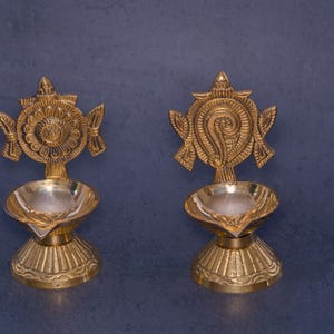 以下が含まれることがあります： 精巧な花柄と渦巻き模様が施された、2つの真鍮製オイルランプ。各ランプにはオイル用の小さなボウルと装飾的な上部があります。ランプは金色で、濃い青色の背景に、テクスチャ加工されたベースの上に立っています。