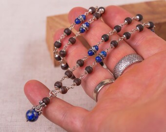 May include: A handcrafted necklace with dark brown wooden beads, silver-toned accents, and blue beads. The necklace has a silver chain and a blue lapis lazuli pendant. The jewelry is held in a hand.