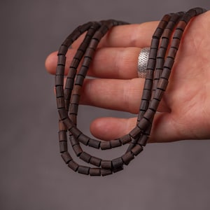 Könnte beinhalten: Drei Stränge dunkelbrauner Holzperlenketten werden in einer Hand gehalten. Die Perlen sind zylindrisch und gleichmäßig verteilt. Der Hintergrund ist einfarbiges, neutrales Grau.