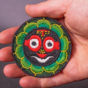 Op de afbeelding: Ronde geborduurde patch met een gestileerd gezicht. Het gezicht heeft grote witte ogen, een rood bovenstuk en een witte glimlach, tegen een zwarte achtergrond. Het is omlijst door groene en gele bladachtige vormen.