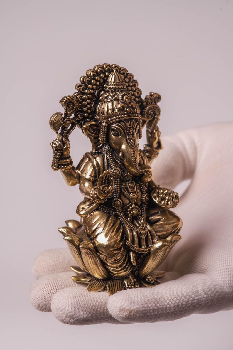 May include: A detailed brass statue of the Hindu deity Ganesha, seated on a lotus flower. The sculpture features intricate details, including ornate jewelry and a large headdress. The statue is held in a gloved hand.