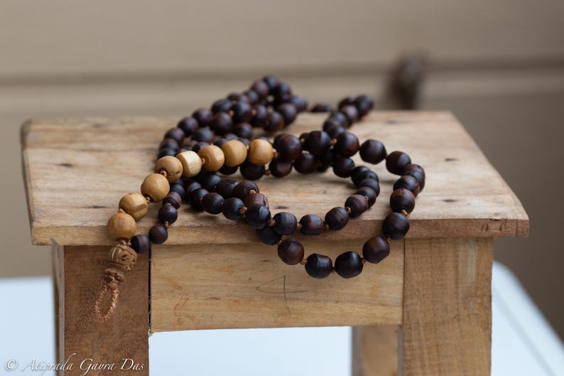 May include: Two wooden prayer bead necklaces rest on a small, rustic wooden stool. One necklace features dark brown beads, while the other has a mix of light and dark brown beads. The beads are strung on a cord.