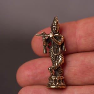 May include: A small, detailed brass figurine of a deity playing a flute. The figure is standing on a lotus flower base, with intricate clothing and jewelry. The statue is held in a hand against a neutral background.