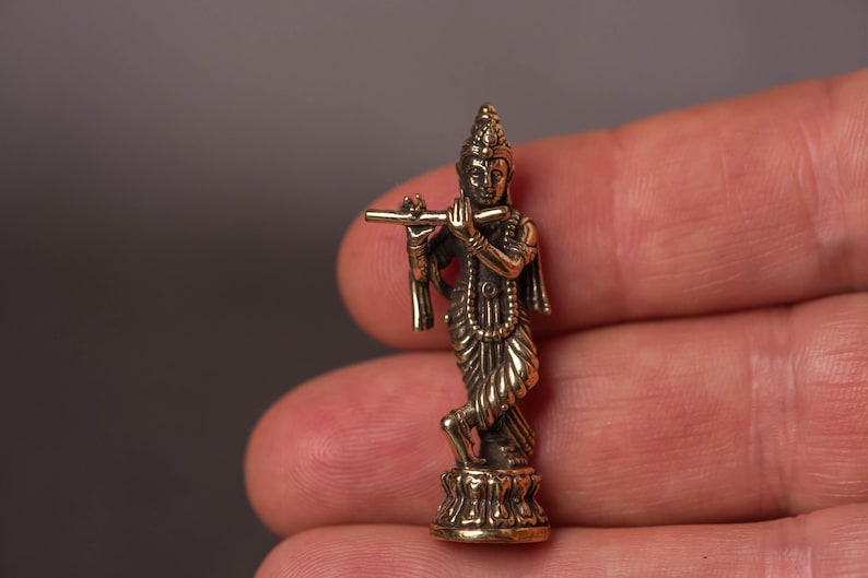 May include: A small, detailed brass figurine of a deity playing a flute. The figure is standing on a lotus flower base, with intricate clothing and jewelry. The statue is held in a hand against a neutral background.