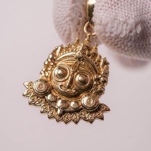 May include: A gold pendant featuring a detailed, embossed face with large circular eyes, a central forehead marking, and an ornate crown. The pendant has a polished metallic finish and includes a small circular bail at the top for attachment.
