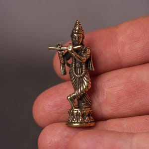 May include: A small, detailed brass figurine of a deity playing a flute. The figure is in a standing pose, with intricate details in the clothing and ornamentation. The figurine is held in a hand, showcasing its size and craftsmanship.