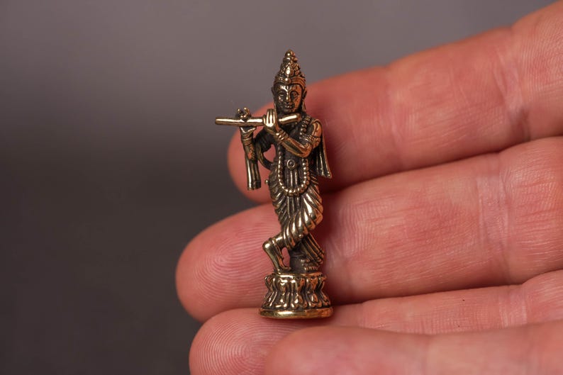 May include: A small, detailed brass figurine of a deity playing a flute. The figure is in a standing pose, with intricate details in the clothing and ornamentation. The figurine is held in a hand, showcasing its size and craftsmanship.