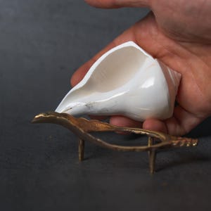 May include: A white seashell is held above a small, ornate, gold-colored metal stand. The stand has a curved design and three short legs. The background is a dark gray surface.