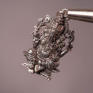 May include: A detailed silver pendant depicting the Hindu deity Ganesha. The pendant is intricately designed with ornate details and is held by a tool. The pendant is a religious item.