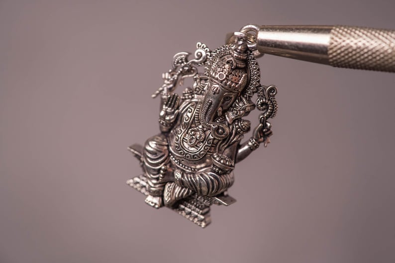 May include: A detailed silver pendant depicting the Hindu deity Ganesha. The pendant is intricately designed with ornate details and is held by a tool. The pendant is a religious item.