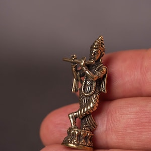 May include: A small, detailed brass figurine of a deity playing a flute. The figure is standing on a decorative base and is adorned with intricate jewelry and a headdress. The figurine is held in a hand against a neutral background.