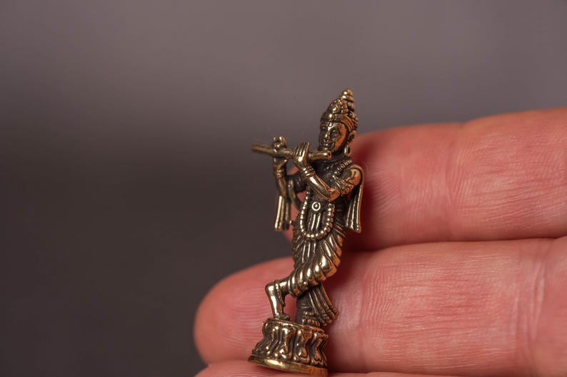 May include: A small, detailed brass figurine of a deity playing a flute. The figure is standing on a decorative base and is adorned with intricate jewelry and a headdress. The figurine is held in a hand against a neutral background.