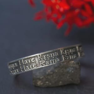Könnte beinhalten: Ein silberfarbener Metallring mit zwei parallelen Bändern, die mit dem Text Hare Krsna Krsna und Hare Rama Rama graviert sind. Der Ring hat eine verwitterte, strukturierte Oberfläche und ist horizontal vor einem dunklen Hintergrund platziert.