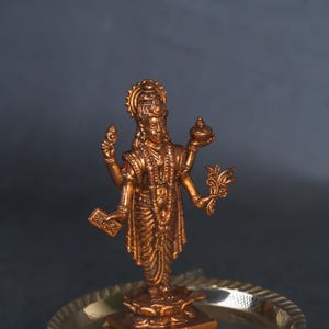 Peut inclure: Statue de divinité de couleur bronze, debout sur une base de lotus, tenant divers objets dans ses multiples bras. La statue est placée sur un plateau métallique rond et décoratif. Le fond est gris foncé.