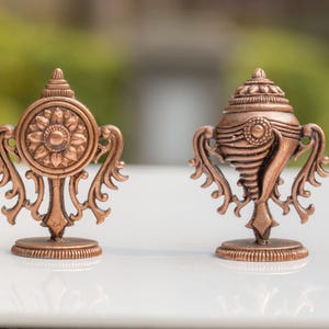 Peut inclure: Deux sculptures décoratives de couleur bronze. L'une présente un motif circulaire avec un centre floral et des accents courbes ornés. L'autre a la forme d'un coquillage avec des éléments décoratifs similaires. Les deux sont sur des bases rondes.