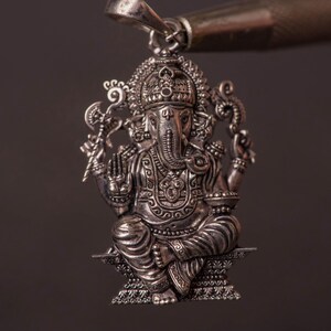 May include: A detailed silver pendant depicting the Hindu deity Ganesha. The pendant features intricate carvings of the elephant-headed god in a seated pose, with multiple arms and ornate details. The pendant is suspended from a silver bail.