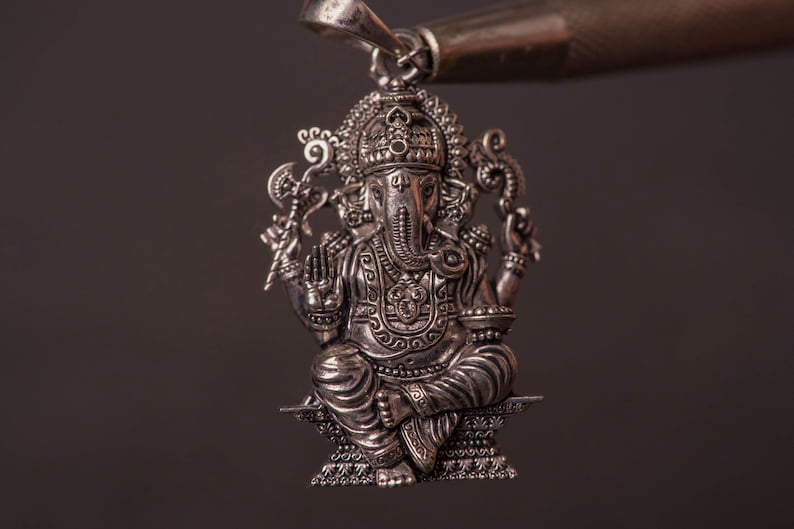 May include: A detailed silver pendant depicting the Hindu deity Ganesha. The pendant features intricate carvings of the elephant-headed god in a seated pose, with multiple arms and ornate details. The pendant is suspended from a silver bail.
