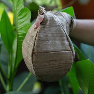 Puede incluir: Una bolsa beige en forma de lágrima con cierre de cremallera, sostenida contra un fondo de follaje verde. La bolsa tiene una superficie texturizada y un pequeño lazo de metal en la parte superior. La bolsa está hecha de tela de aspecto natural.