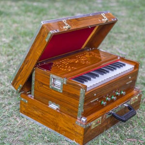 Pode incluir: Um harmônio de madeira com uma tampa articulada, aberta para revelar um interior vermelho. O instrumento possui um teclado com teclas pretas e brancas e vários botões verdes. A caixa de madeira é decorada com detalhes metálicos e um desenho floral.