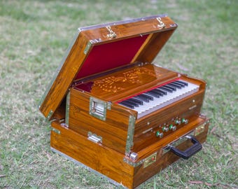 May include: A wooden harmonium with a hinged lid, open to reveal a red interior. The instrument has a keyboard with black and white keys, and several green knobs. The wooden case is decorated with metal accents and a floral design.