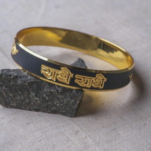 May include: Gold-toned bangle with a black enamel band. The bangle features gold lettering and decorative accents. The jewelry is displayed on a dark gray rock against a neutral fabric background. The text on the bangle reads "राधे राधे".