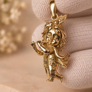 Könnte beinhalten: Ein Anhänger aus poliertem Gold in Form einer flötenspielenden Figur. Die Figur hat detailliertes Haar mit einem Pfauenfeder-Akzent, trägt traditionelle drapierte Kleidung und hält eine horizontale Flöte an die Lippen. Oben befindet sich eine Öse.