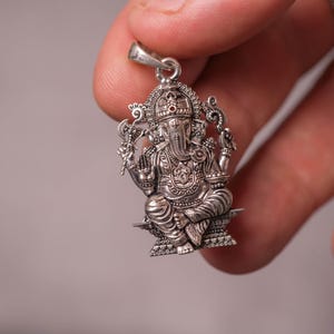 May include: A detailed silver pendant featuring the Hindu deity Ganesha, seated in a cross-legged pose. The pendant showcases intricate carvings, a small red stone, and a bail for a chain. The design includes ornate details on the crown and clothing.