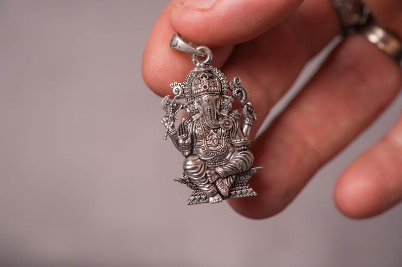 May include: A detailed silver pendant featuring the Hindu deity Ganesha, seated in a cross-legged pose. The pendant showcases intricate carvings, a small red stone, and a bail for a chain. The design includes ornate details on the crown and clothing.