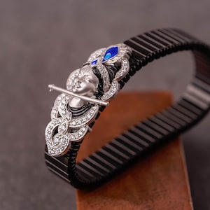 Puede incluir: Una pulsera negra con una pieza central plateada detallada. La pieza central presenta una figura tocando la flauta, adornada con piedras preciosas transparentes y detalles azules. La pulsera está hecha de eslabones negros.