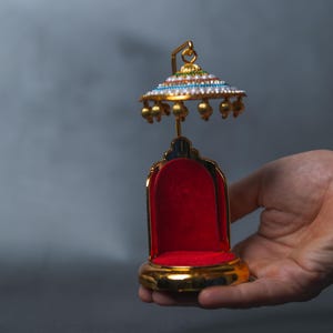 Peut inclure: Un petit trône doré avec un siège en velours rouge et un auvent décoratif. L'auvent est orné de bijoux colorés et d'accents dorés. Le trône est tenu dans la main d'une personne sur un fond gris.