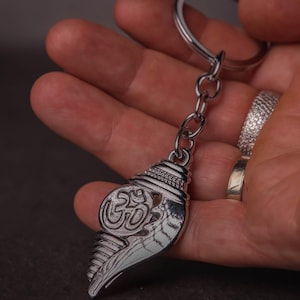 Puede incluir: Llavero plateado con un diseño de concha con el símbolo Om. El llavero está unido a una cadena y un anillo plateados. La concha tiene crestas detalladas y un acabado pulido.