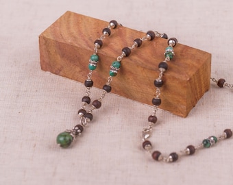 May include: A necklace with a silver chain, dark brown wooden beads, and turquoise accents. The necklace is displayed on a wooden block. The pendant is a green stone. The design highlights the natural materials and craftsmanship.