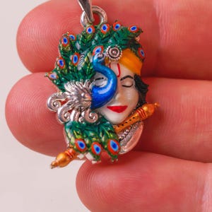 Könnte beinhalten: Silberanhänger mit einer farbenfrohen Darstellung einer Gottheit mit Pfau und Flöte. Der Anhänger hat ein detailliertes Design mit grünen, blauen, gelben und roten Akzenten. Der Anhänger wird von einer Person gehalten.