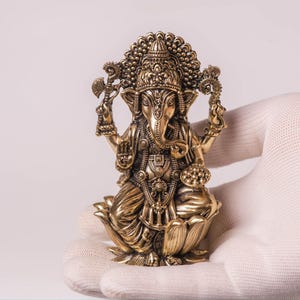 May include: A detailed brass statue of the Hindu deity Ganesha, seated on a lotus flower. The statue features intricate carvings, a large head, and multiple arms holding various objects. The figure is held in a white gloved hand against a plain background.