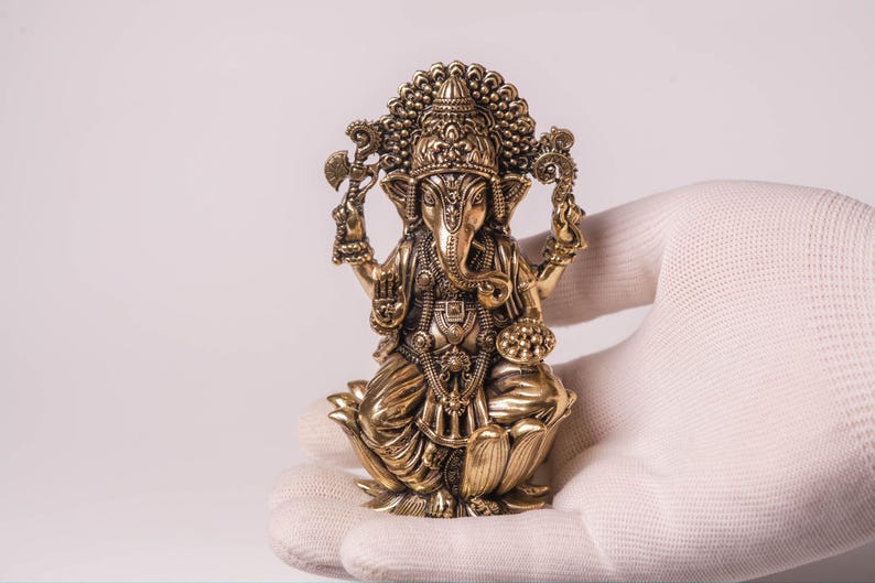 May include: A detailed brass statue of the Hindu deity Ganesha, seated on a lotus flower. The statue features intricate carvings, a large head, and multiple arms holding various objects. The figure is held in a white gloved hand against a plain background.